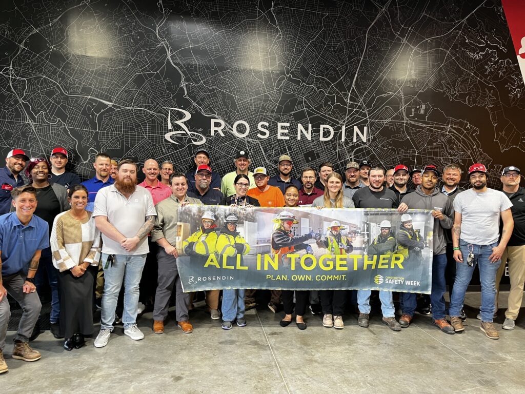 A large group of Rosendin employees stands together, proudly holding a banner that reads "All in together" in celebration of Rosendin Safety Week